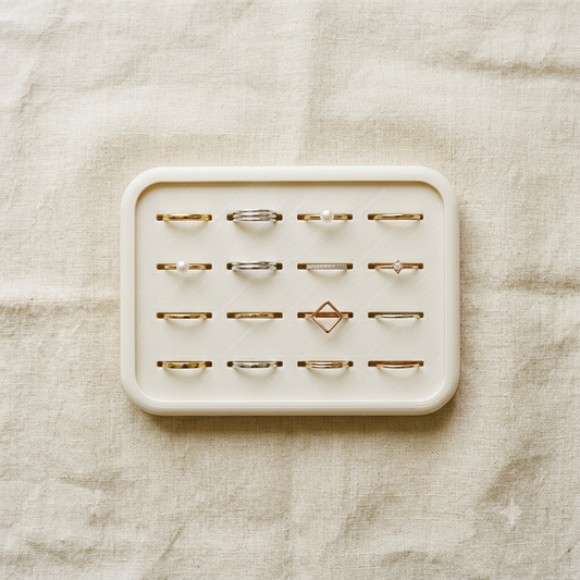 A cream-colored rectangular ring display tray holding 15 minimalist rings in silver, gold, and rose gold tones, arranged on a textured beige linen fabric background.